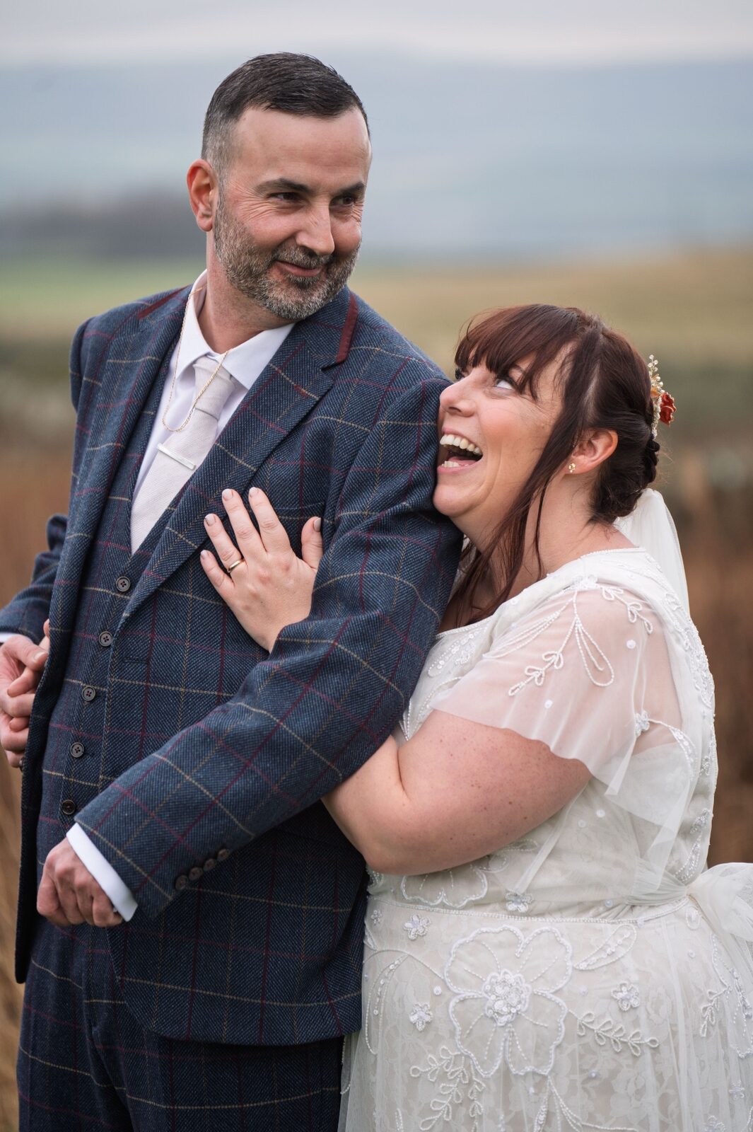 A bride laughs warmly against her groom's shoulder, both lit by soft winter light on a hillside.