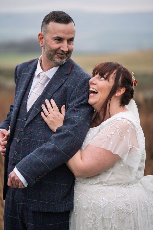 A bride laughs against the shoulder of her groom in a tweed three-piece suit, with rolling hills behind them.