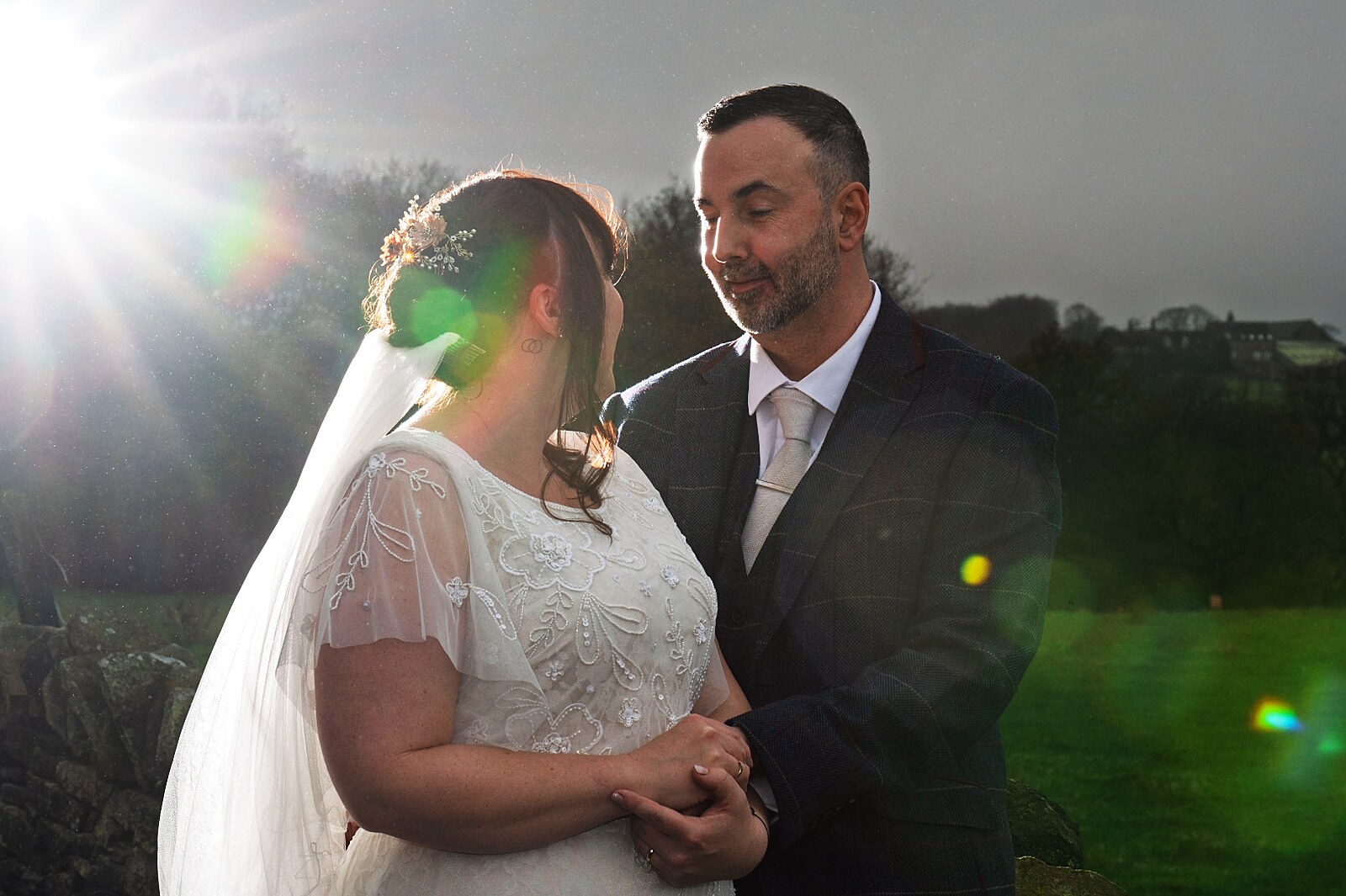 A bride and groom share a quiet moment outdoors, backlit by a low winter sun that bursts across the frame in a soft flare.