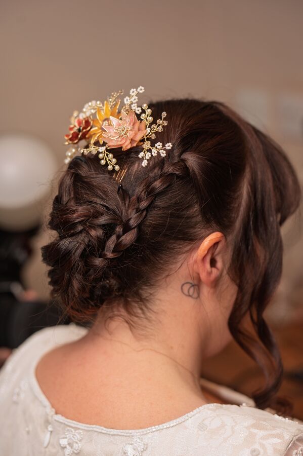 The back of a bride's plaited updo finished with a delicate floral and pearl hairpiece in warm tones.