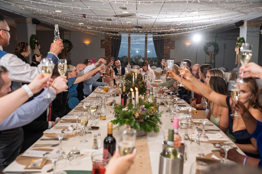 Wedding guests stand at a long candle-lit table beneath fairy lights, raising champagne flutes towards the couple.